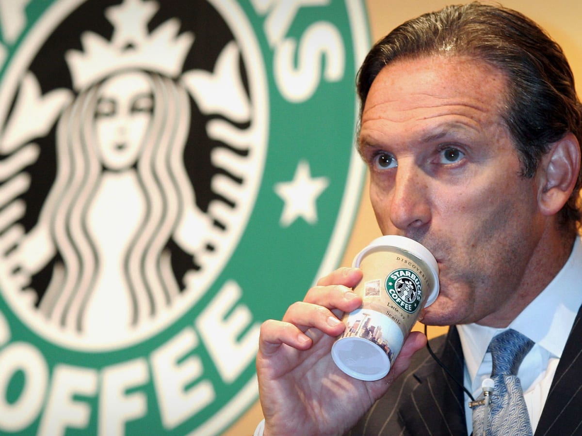 Schutz tomando café em um copo da Starbucks, vestindo terno e com a logo da companhia ao fundo.