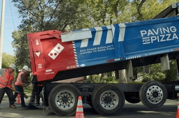 Caminhão da campanha "pavimentando por pizzas" e 3 trabalhadores asfaltando uma rua.