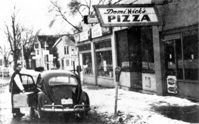 Foto preta e branca da fachada da Dominicks Pizza, com um homem e um fusca em frente ao estabelecimento.