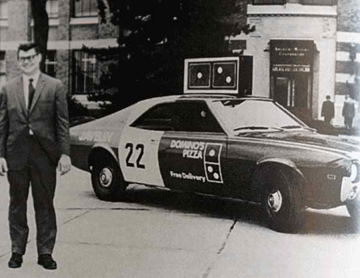 Foto preta e branca com um rapaz à frente e um carro de entregas da Dominos Pizza ao fundo.