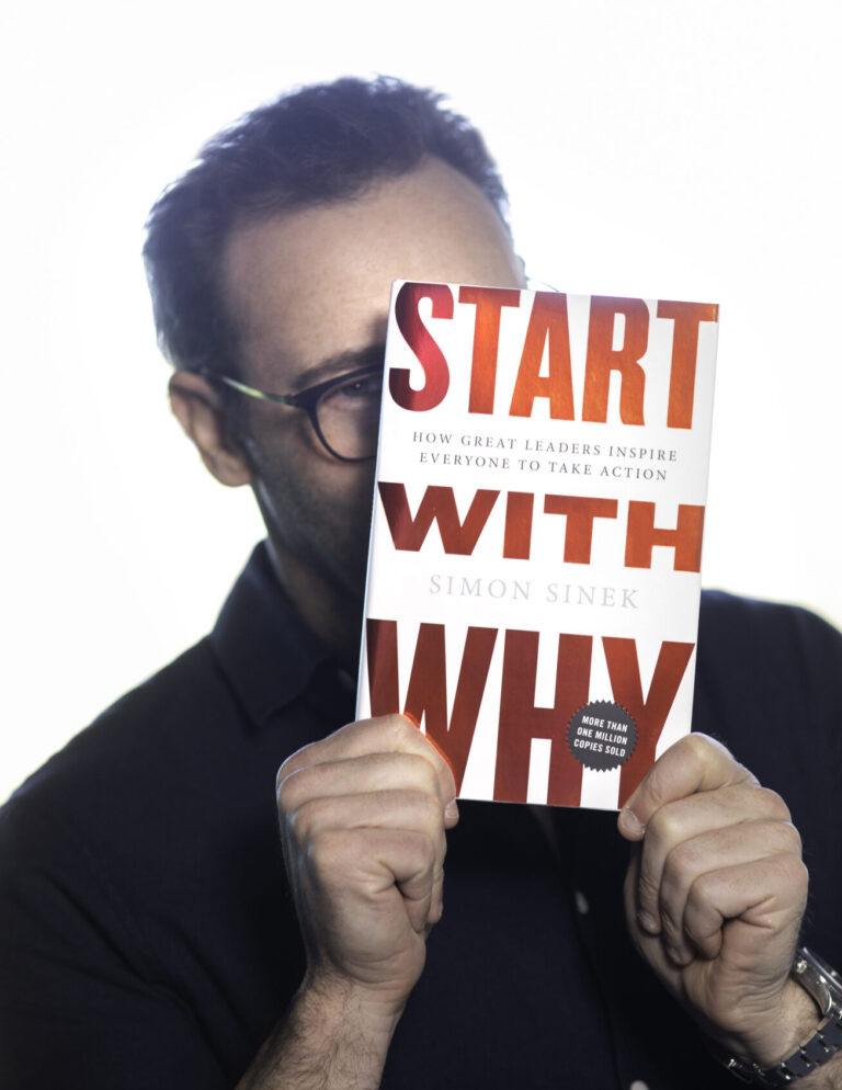 Simon Sinek de camisa preta e óculos, tampando o rosto e mostrando a capa do seu livro "Comece pelo porquê".